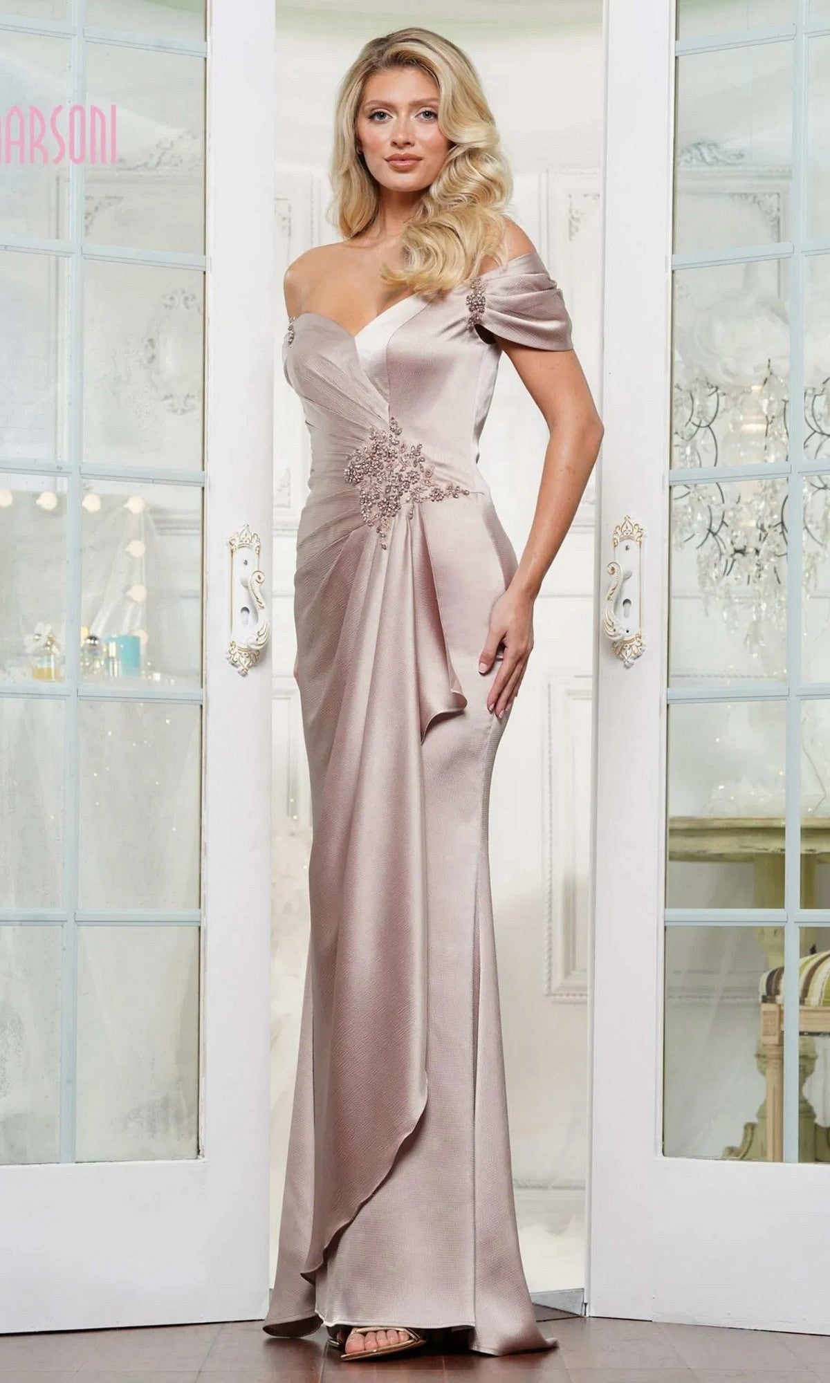 A woman stands indoors wearing the Marsoni MV1351 long formal dress in sand, an off-the-shoulder, floor-length evening gown with floral embellishments at the waist.